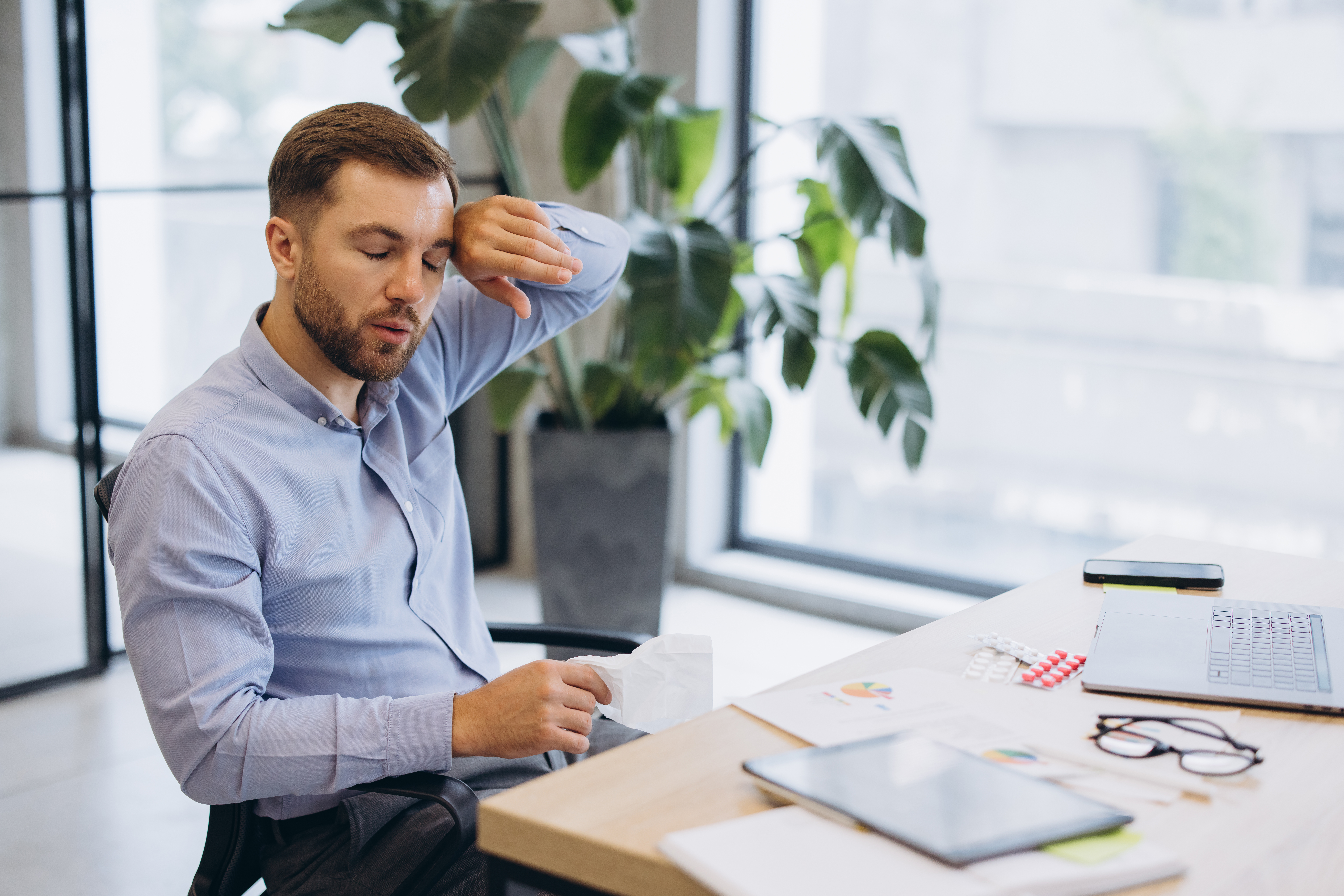 Erkrankter Beschäftigter mit Taschentuch und Unterlagen am Arbeitsplatz